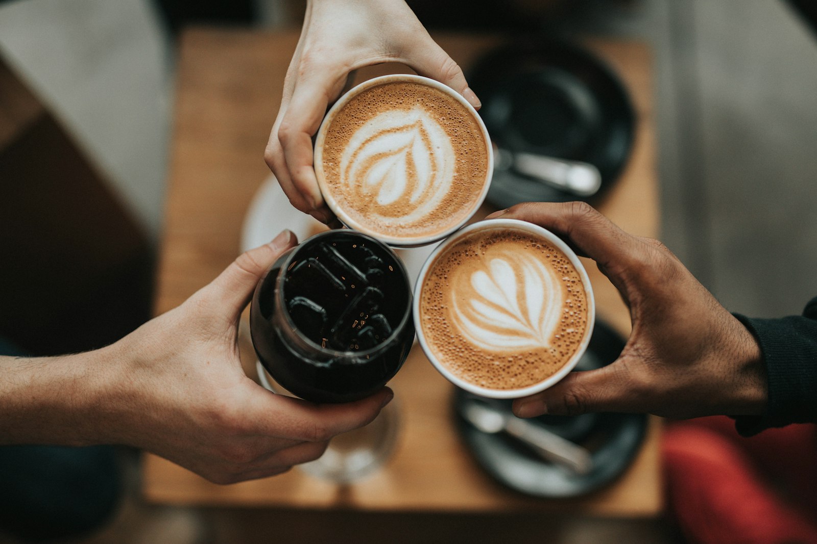 Three hands toasting with two lattes and an iced coffee at a cafe table